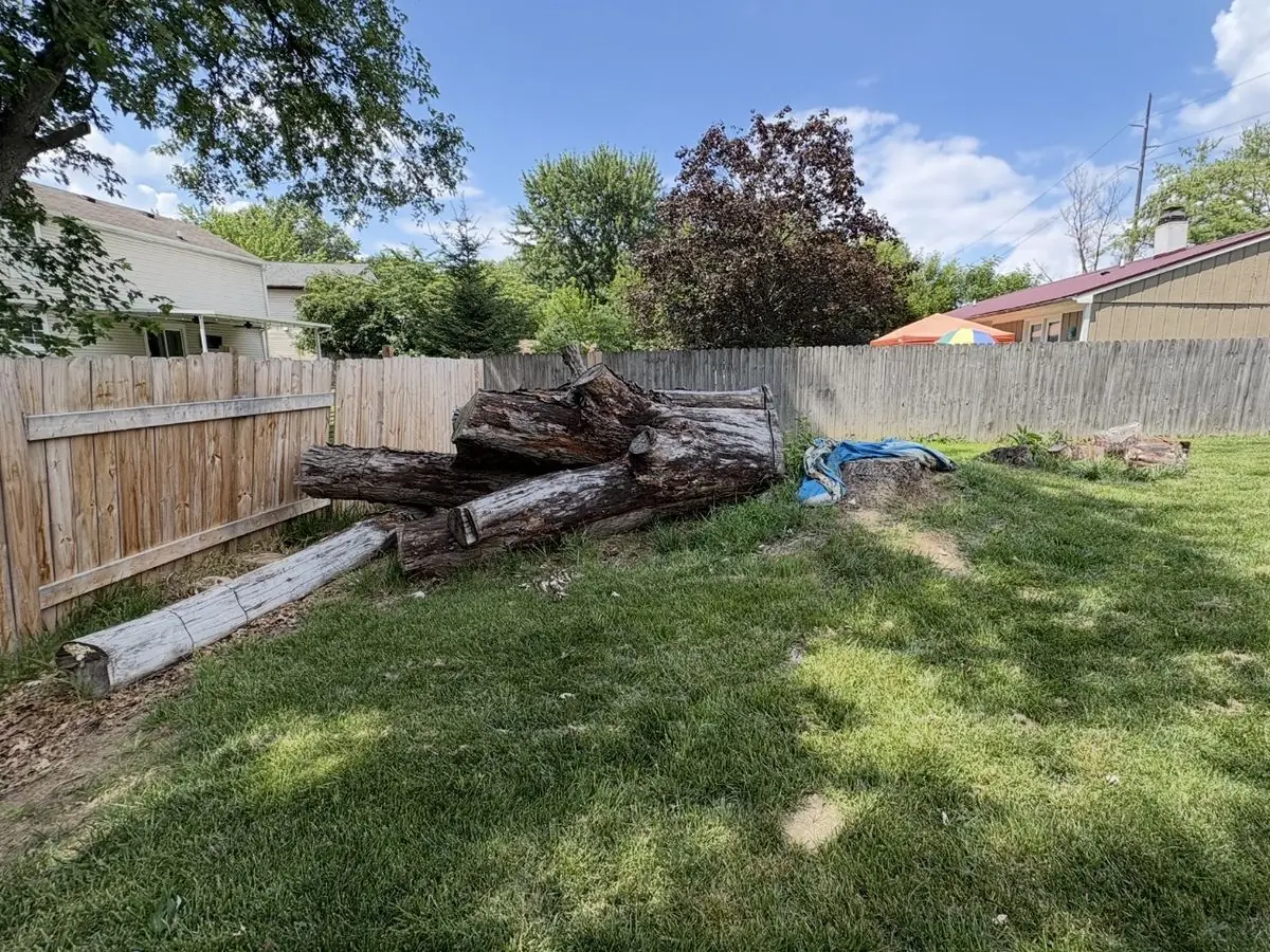 Stack of old logs along a wooden fence and a tree stump in a back yard before cleanup and stump grinding in South Central Indiana