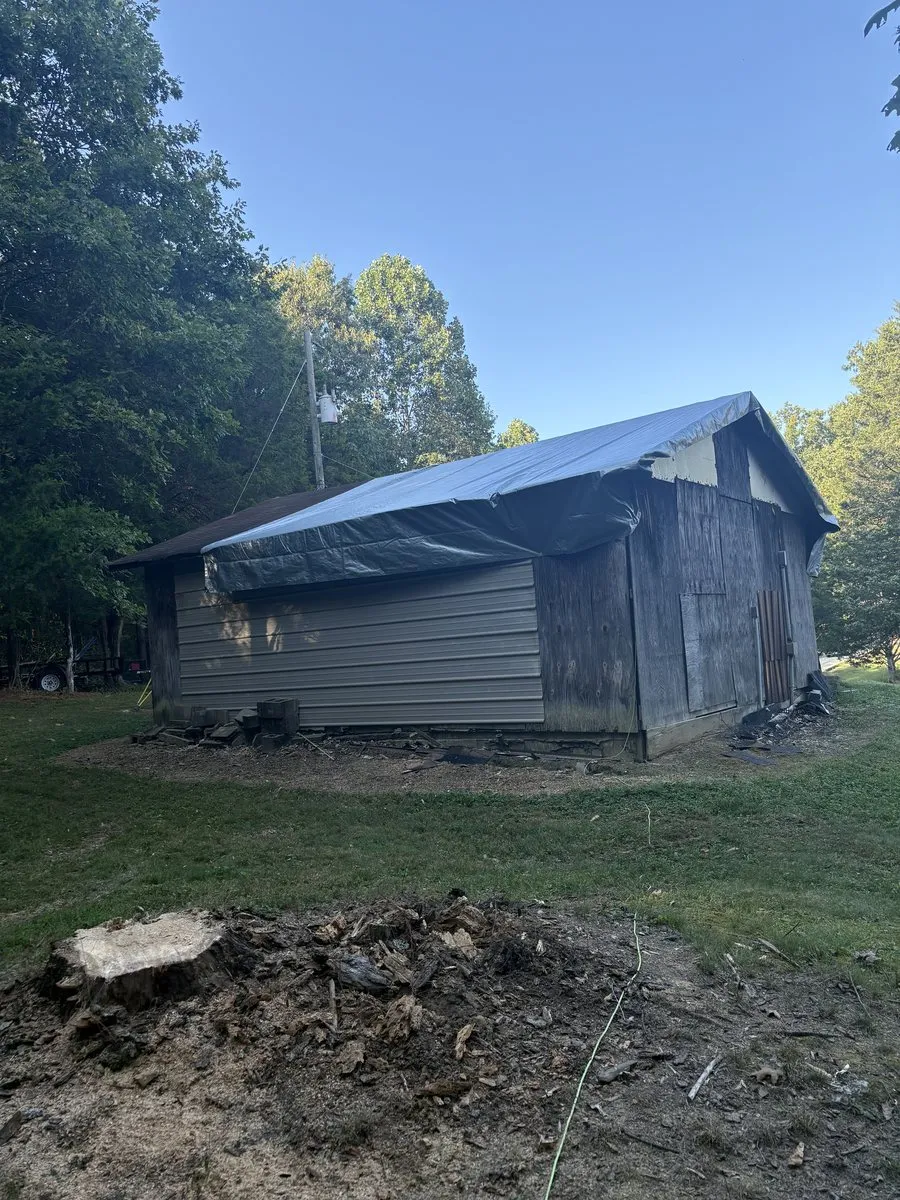 Same garage in Freetown, Indiana after the fallen tree was removed — roof covered with a tarp and the neighboring dead tree cut down to a stump