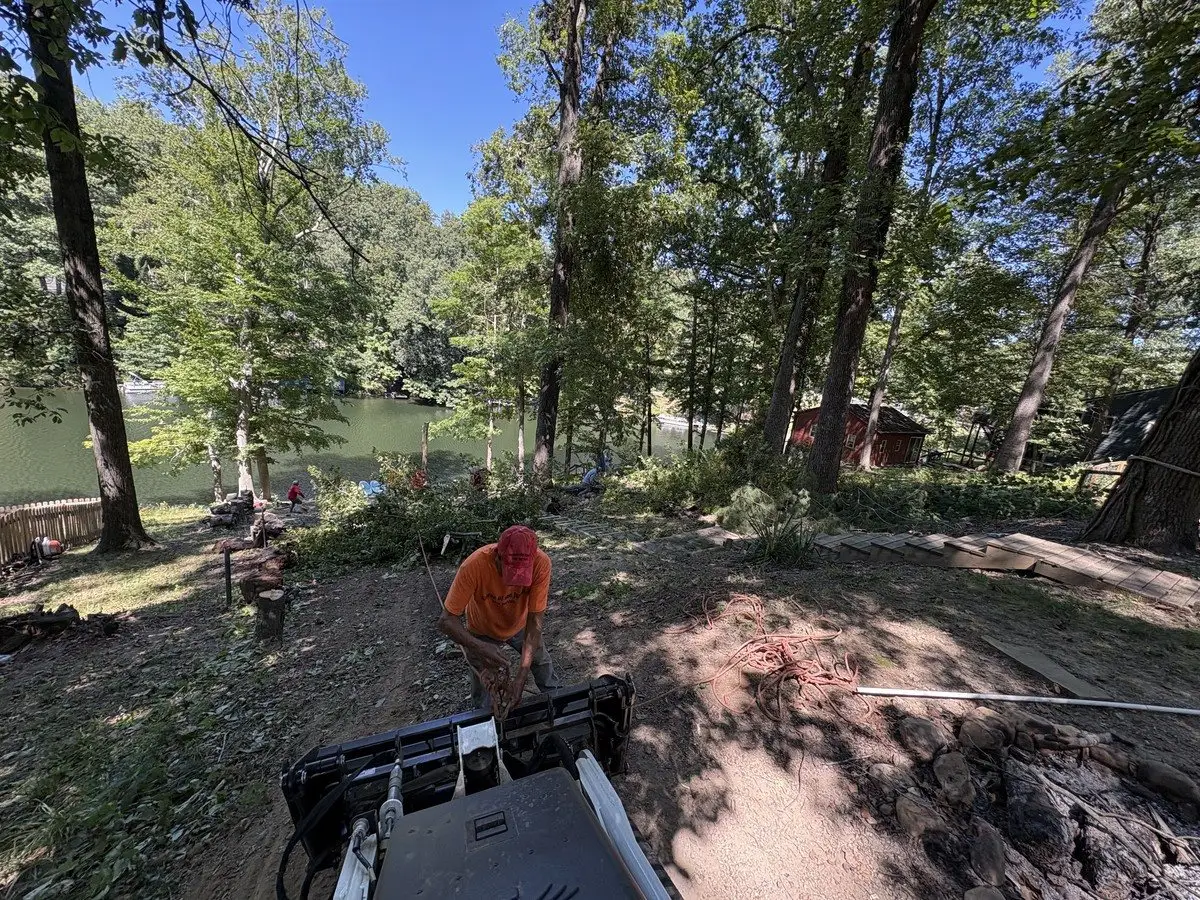 Crew member rigging a rope on a compact loader at the top of a steep bank to haul cut trees up from the lakeshore in Prince's Lakes, Indiana — the lake now showing through the thinned woods