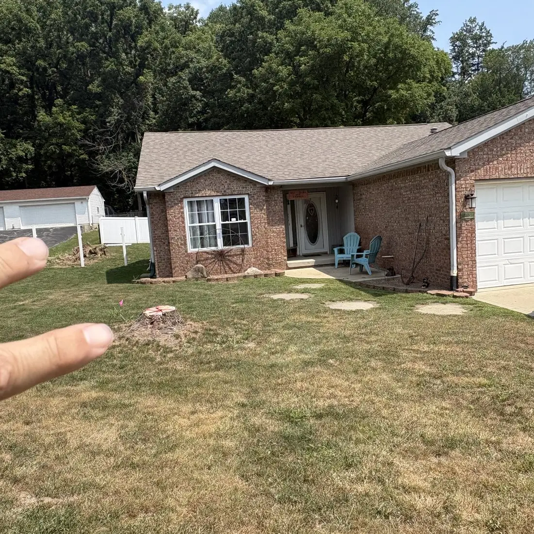 Fresh-cut tree stump in the front lawn of a brick home in Martinsville, Indiana, with the uprooted stump from a July storm visible in the yard behind it, before stump grinding